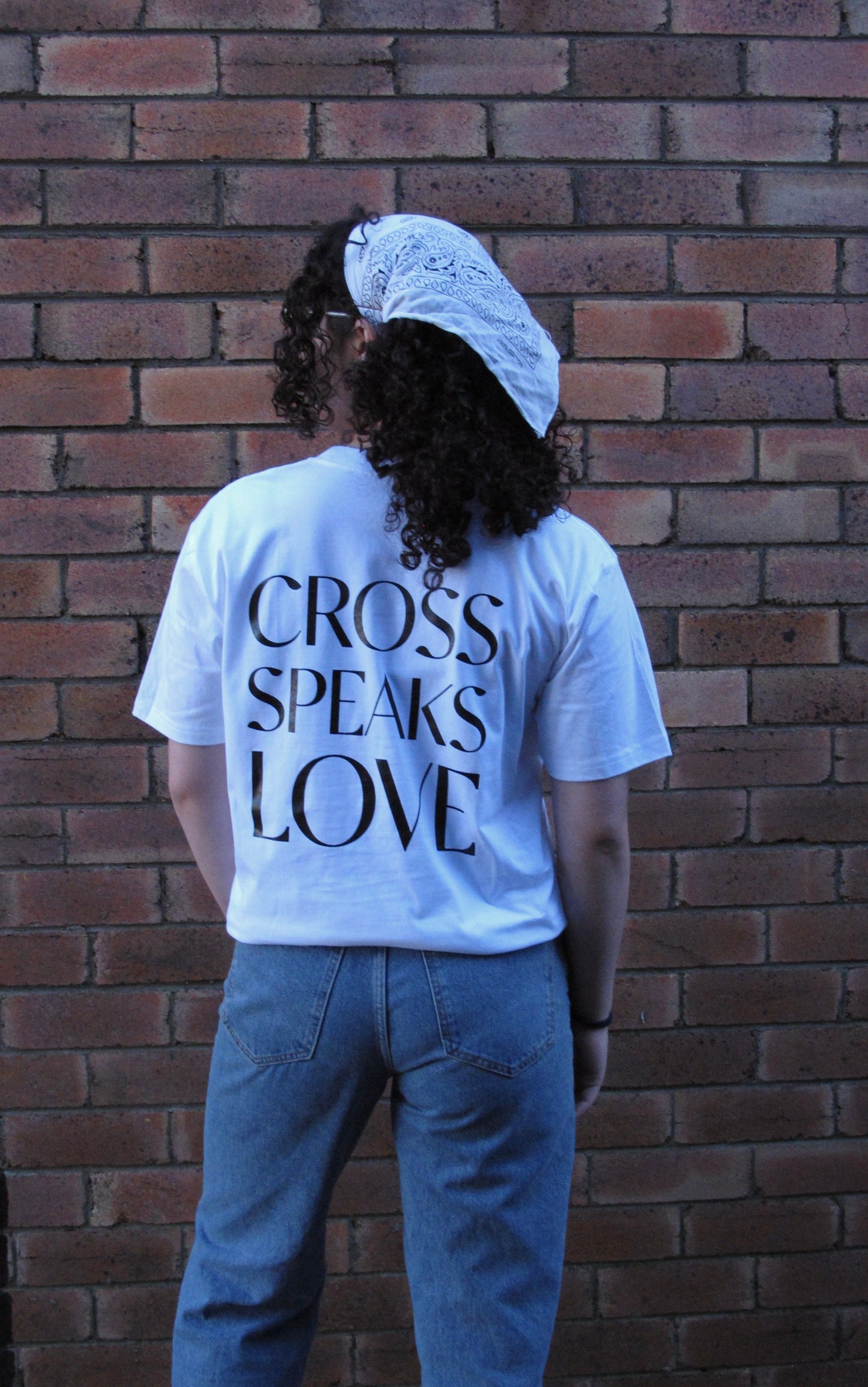 Female modelling with white scarf in curly brown hair. she is facing a brick wall and the words CROSS SPEAKS LOVE is written in bold big print and capitals in black across her back. SHIRT Materials: 100% Australian Cotton, Plastisol Ink Details: -Pocket Print -Back Print -Bottom right SSD print -Straight Hemline -Unisex fit -White T-shirt with Blank ink