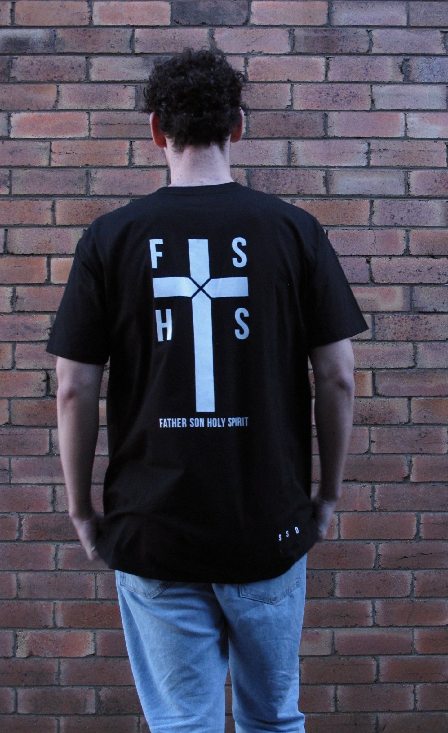 NEW DESIGN - FSHS in the four squares of the cross stating the first letters of the words Father Son Holy Spirit. Design aslo has large cross with rope like design in centre and the words FATHER SON HOLY SPIRIT written in capitals along the base. Right bottom side SSD acronym. All prints in white ink. on black shirt