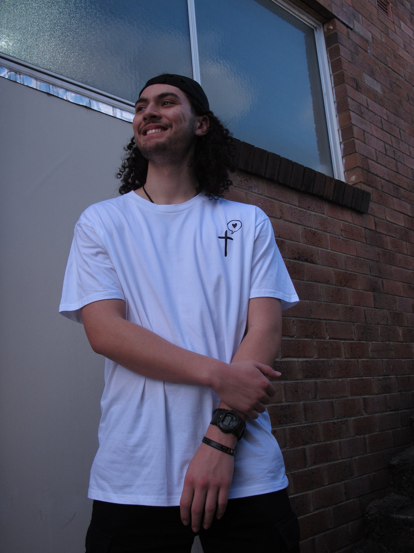 a male wearing a White T-shirt with a pocket print of cross symbol with a speech bubble with a love heart inside printed in black. He wears a Soul Submitted black cap facing backwards. His hair is long and curly and is out. He smiles and crosses his arm casually in front on him