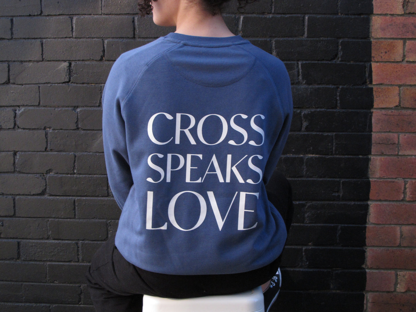 Female model sitting on white stool facing a brick wall painted black. She has her head turned to the left and some of her curls are showing but not her whole back of head. She wears a diesel blue sweatshirt with white wiring CROSS SPEAKS LOVE in capitals as individual lines.