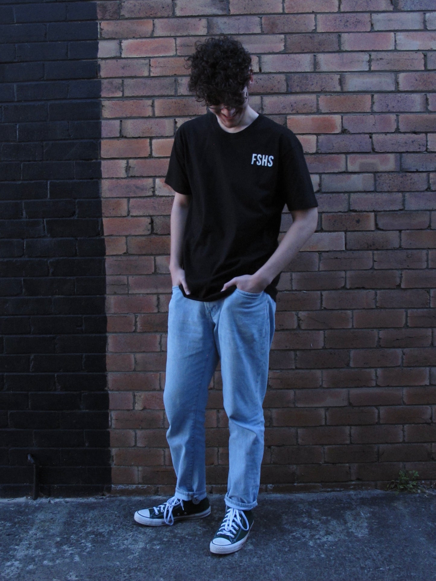 Model looking down standing in front of a brick wall. FSHS pocket print on lefrt side BLACK tee with white ink.