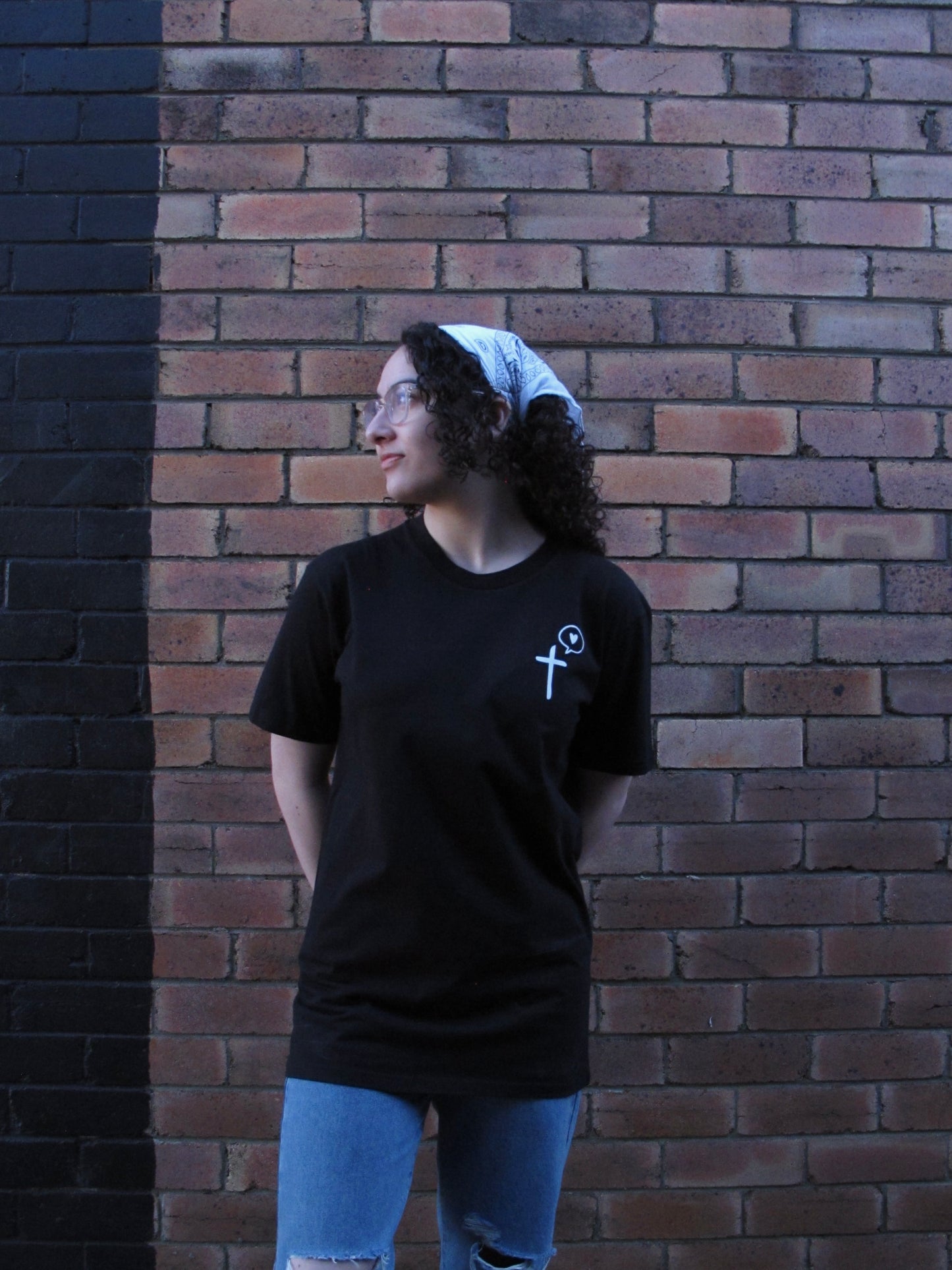 Female model with brown culry hair wearing glasses and white scarf in her hair. Front facing in black tshirt with left side pocket print with image of a cross with a speech bubble of a love eart in it. The design is called - Cross speaks love. The tshirt is untucked and comes down to her top thigh. She accompanies tshirt with blue jeans.