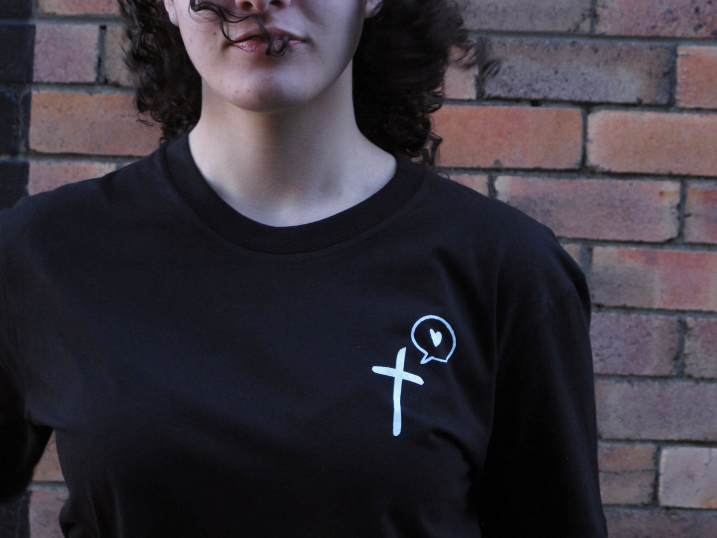 Female model. up close of bottom half of her face and the cross speaks love pocket print design of a cross with a speech bubble to the left with a love heart within it. white ink on black tee . Brick wall begind