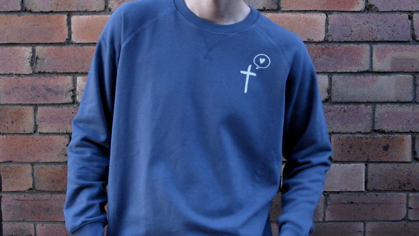Male model leaning up against brick wall wearing diesel blue sweatshirt with cross and speech bublle with love heart in it as pocket print in white ink.
