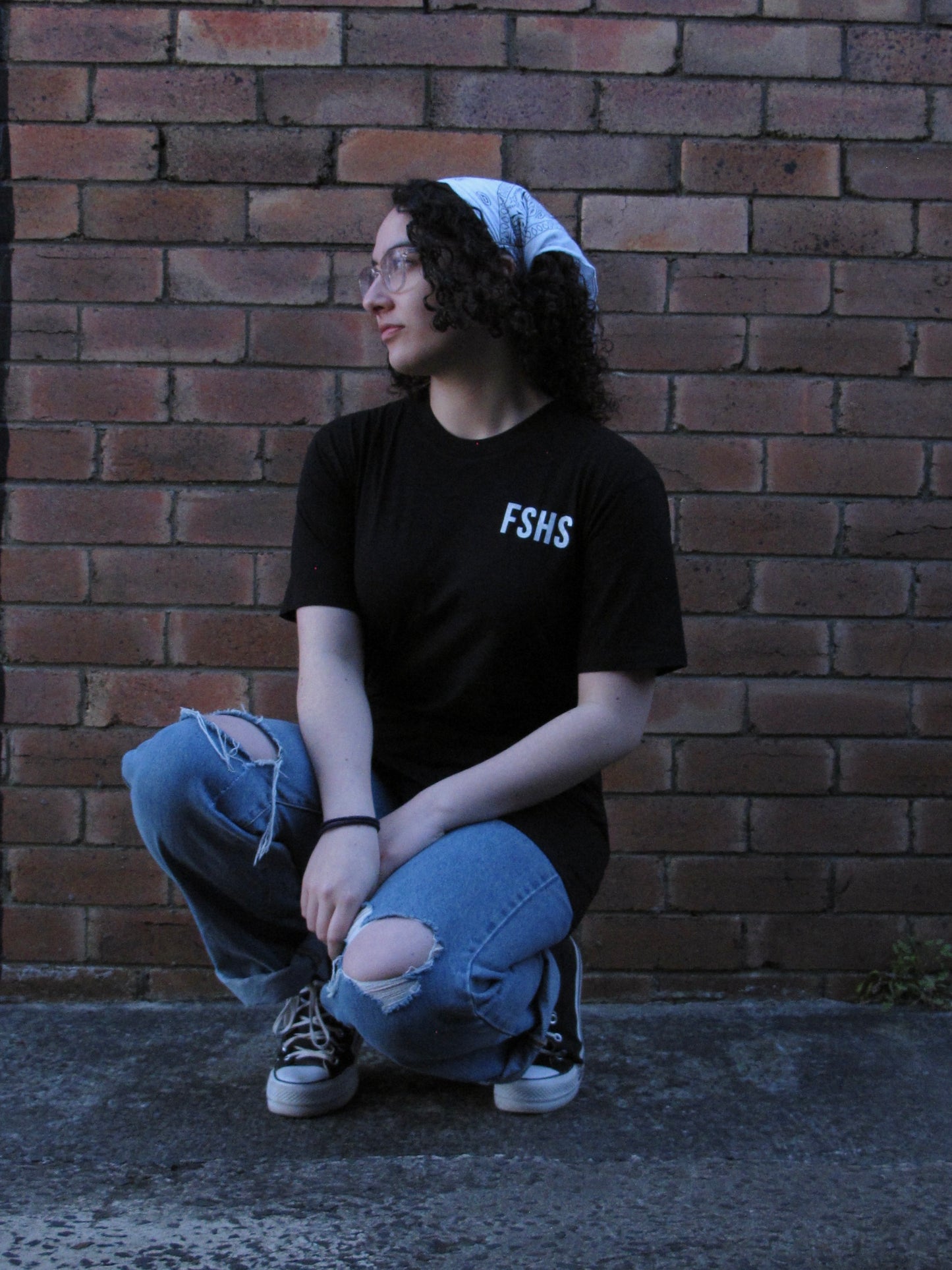 Female model relaxed squating/ kneeling in front of brick wall. She is wearing glasses and a headscarf and our black FSHS tee. This pic has her facing front and pocket print is visible.