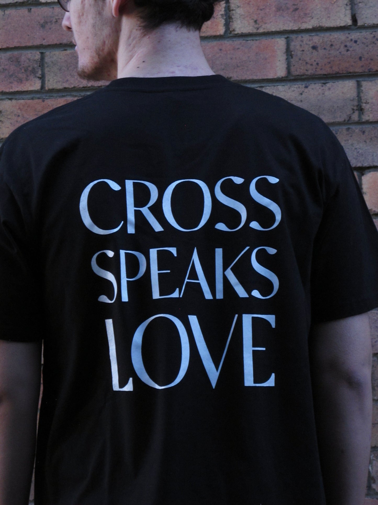 Male model wearing black tshirt with white ink design of words CROSS SPEAKS LOVE in capitals across his mid bacl. His head is turned to the left and a brick wall behind.