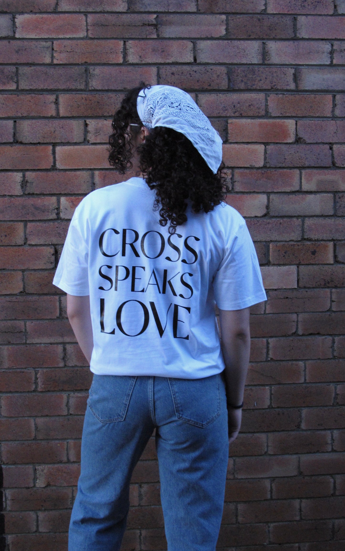 Female modelling with white scarf in curly brown hair. she is facing a brick wall and the words CROSS SPEAKS LOVE is written in bold big print and capitals in black across her back.    SHIRT Materials: 100% Australian Cotton, Plastisol Ink  Details:  -Pocket Print  -Back Print  -Bottom right SSD print  -Straight Hemline  -Unisex fit  -White T-shirt with Blank ink