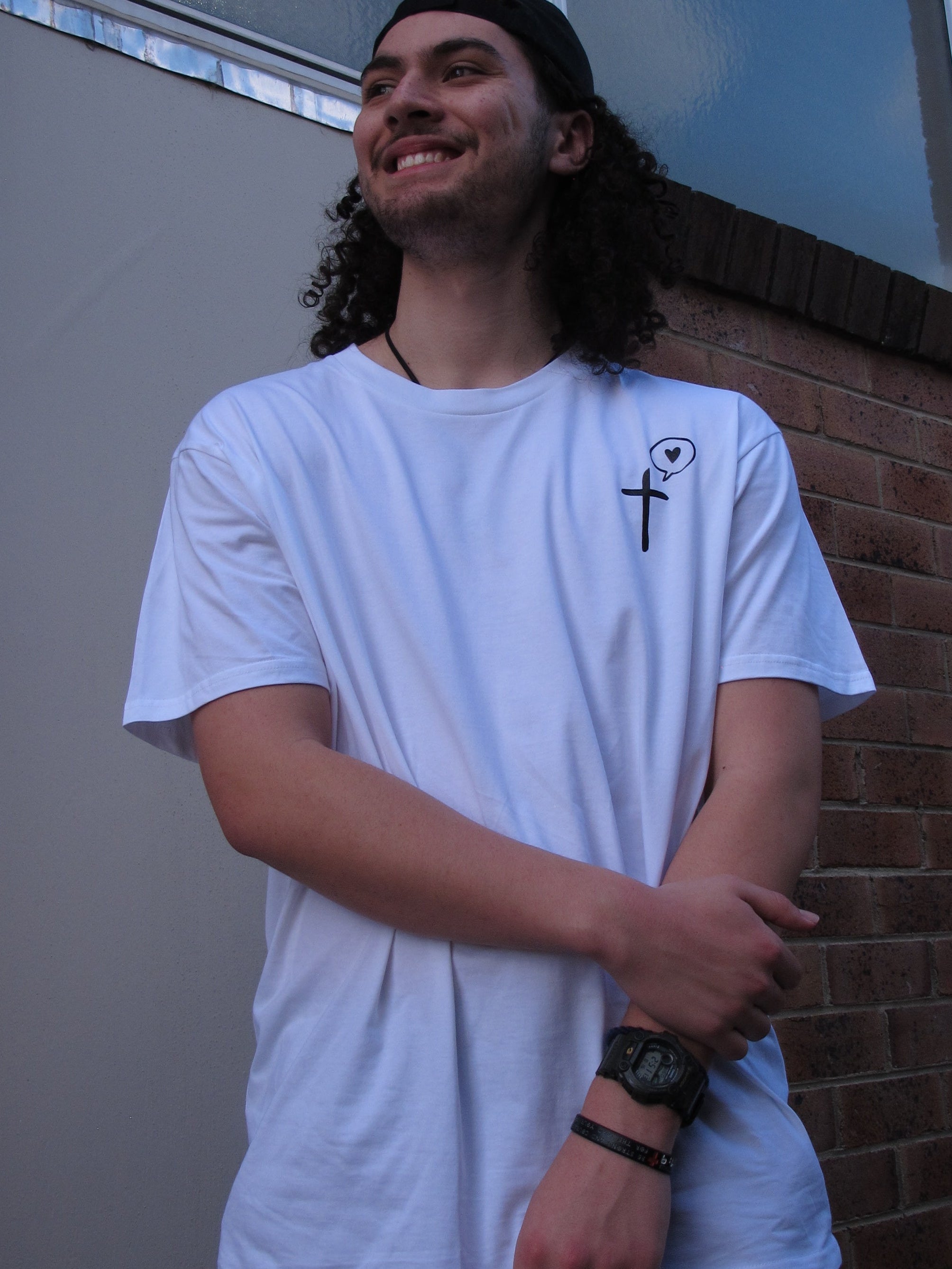 a male wearing a White T-shirt with a pocket print of cross symbol with a speech bubble with a love heart inside printed in black. He wears a Soul Submitted black cap facing backwards. His hair is long and curly and is out. He smiles and crosses his arm casually in front on him 