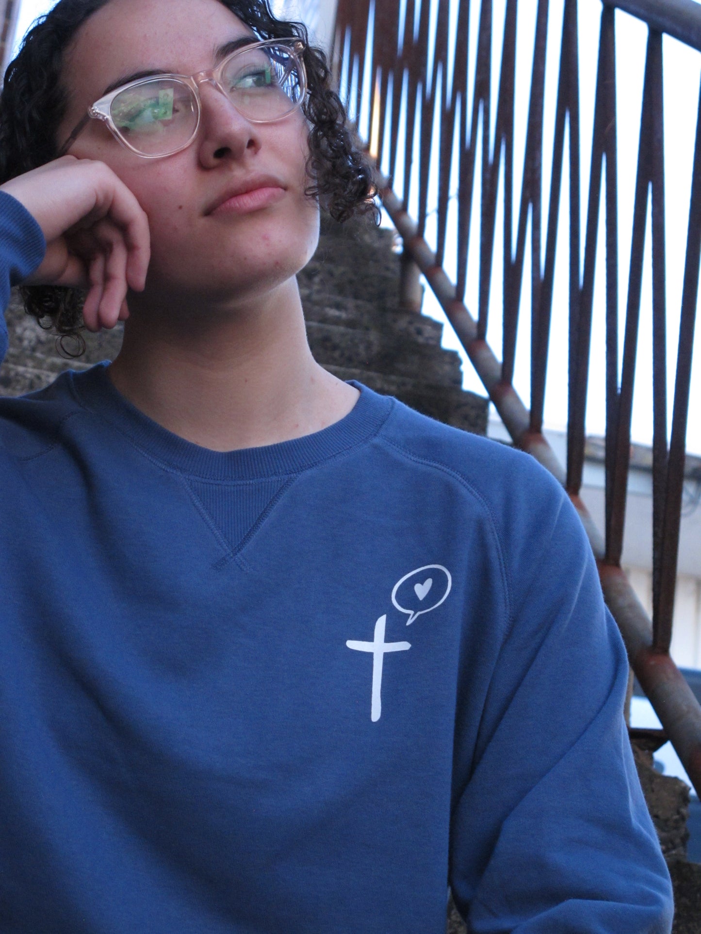 Female resting her head on her hand  - wearing glasses and looking out. She is wearing glasses. Model wears a diesel blue sweatshirt with pocket print on left side. the print is white and is a cross symbol with a speeck bubble with a love heart in it 