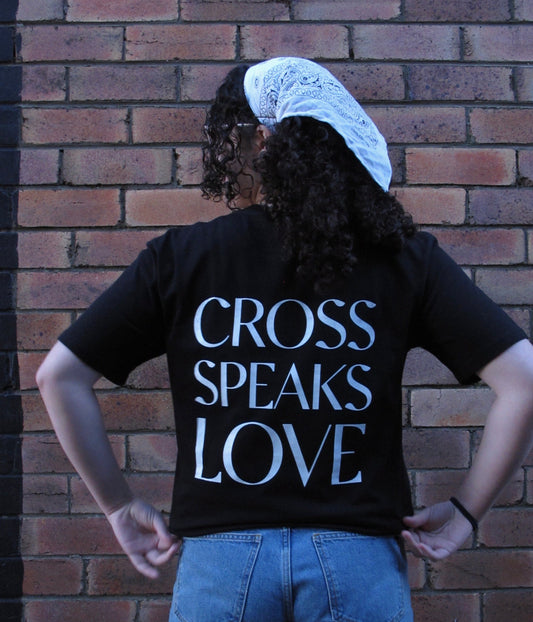 A female wearing a Black T-shirt with an elegant white design on the back Design on the back: CROSS SPEAKS LOVE in capitals as a large bold back print. Model wears a white scarf in her hair and has her hands at her waist pulling on the shirt a little bit