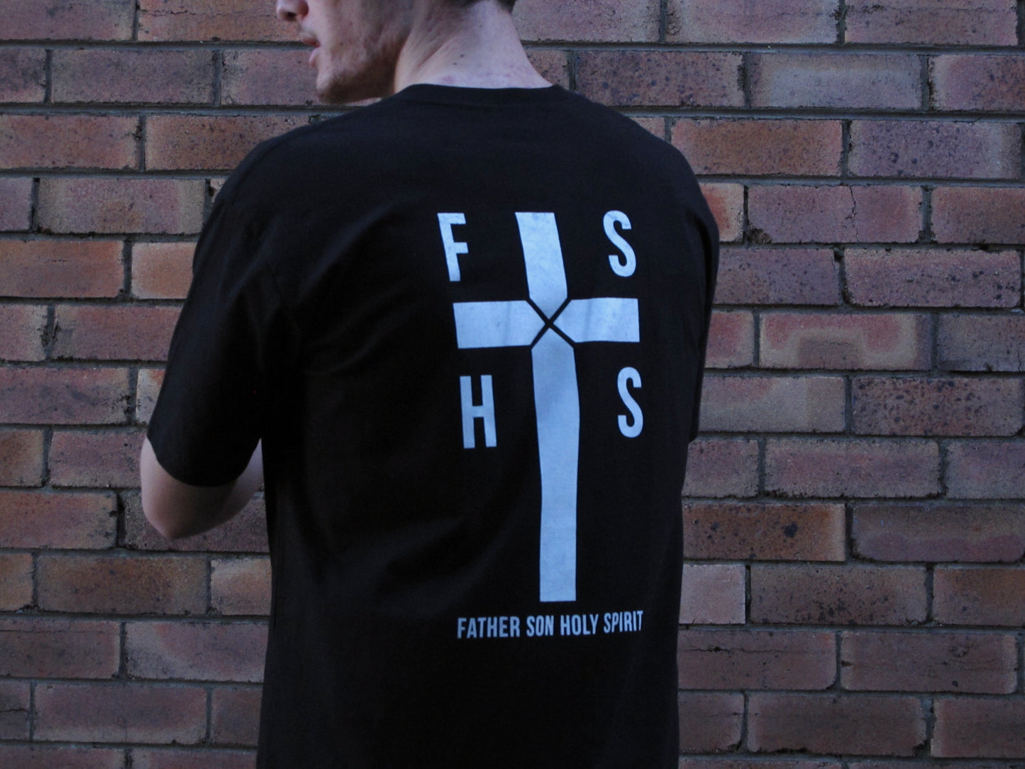 NEW DESIGN - FSHS in the four squares of the cross stating the first letters of the words Father Son Holy Spirit. Design aslo has large cross with rope like design in centre and the words FATHER SON HOLY SPIRIT written in capitals along the base. Right bottom side SSD acronym. All prints in white ink. on black shirt