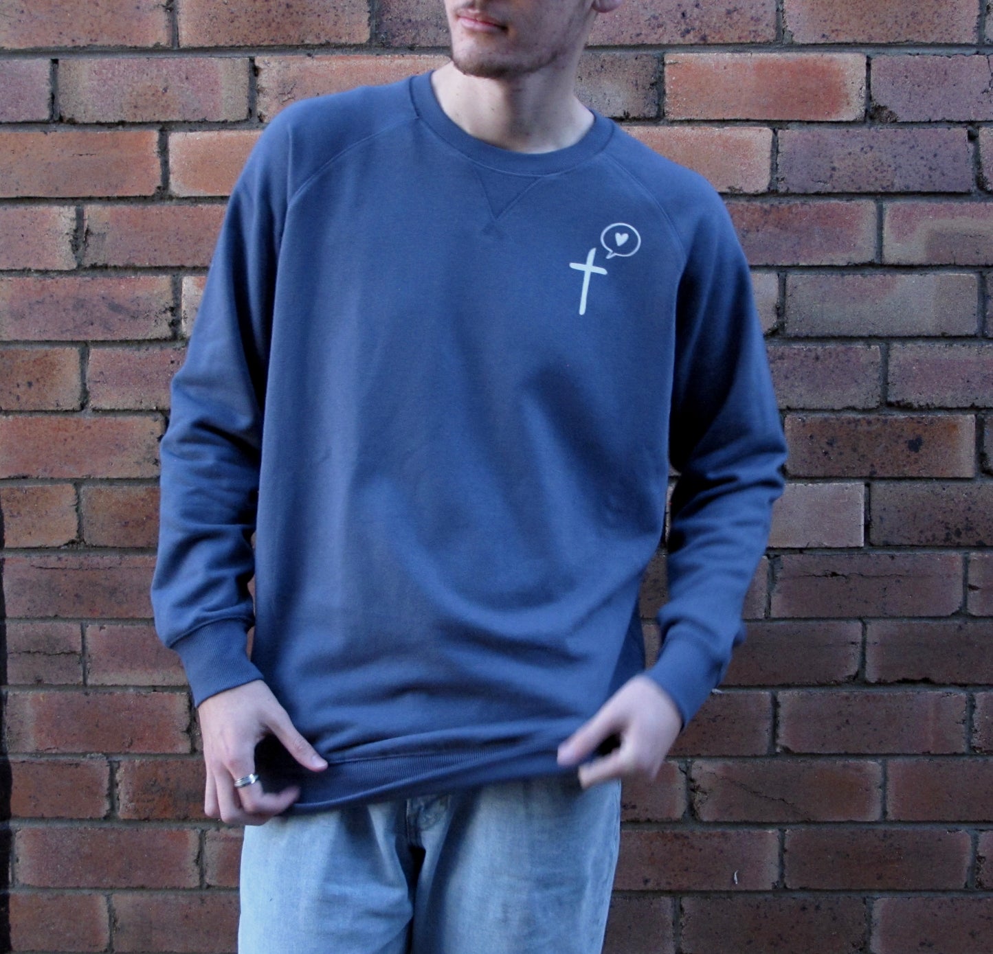 Male model leaning up against brick wall wearing diesel blue sweatshirt with cross and speech bublle with love heart in it as pocket print in white ink. 