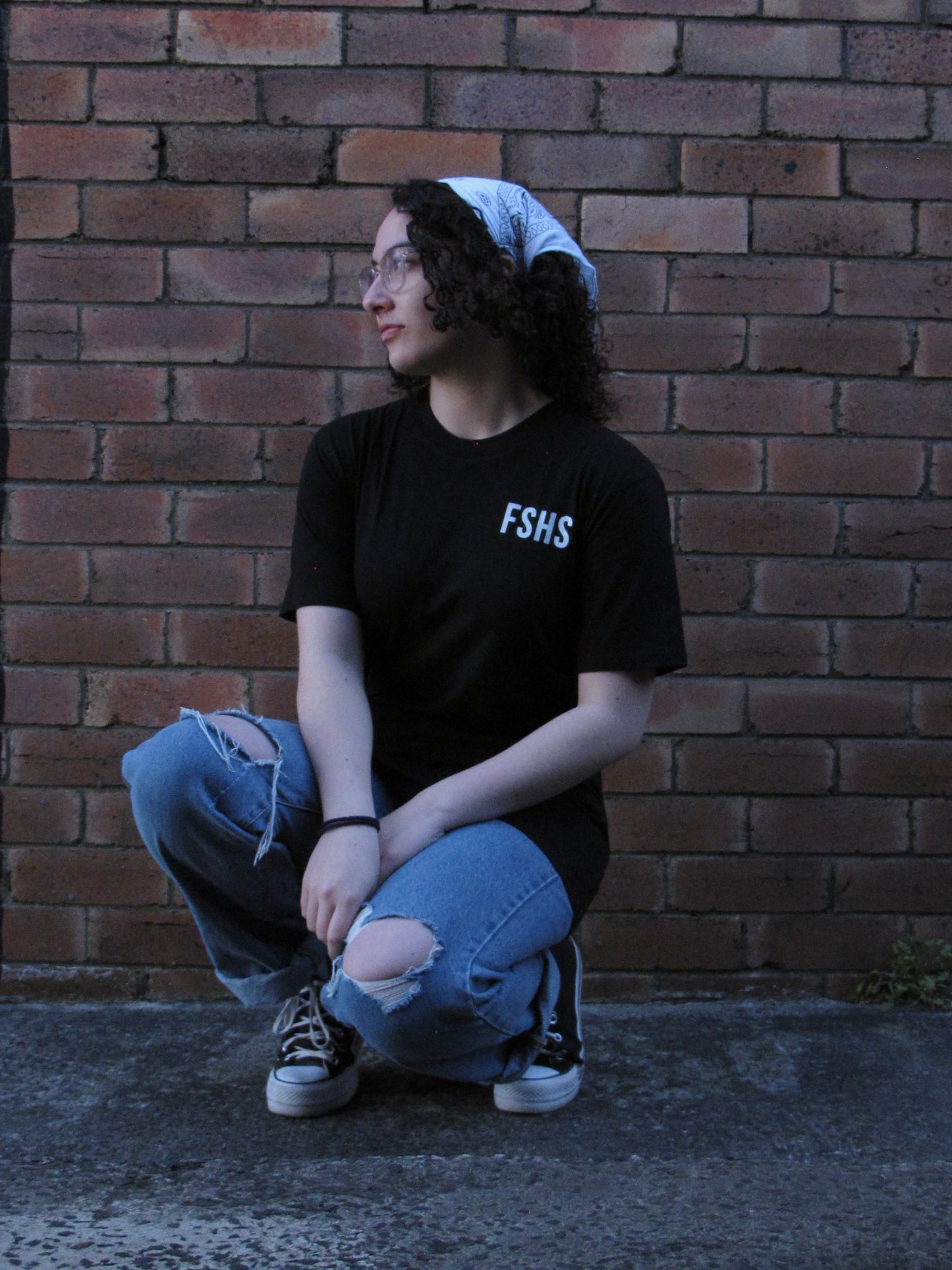 Female model relaxed squating/ kneeling in front of brick wall. She is wearing glasses and a headscarf and our black FSHS tee. This pic has her facing front and pocket print is visible. 