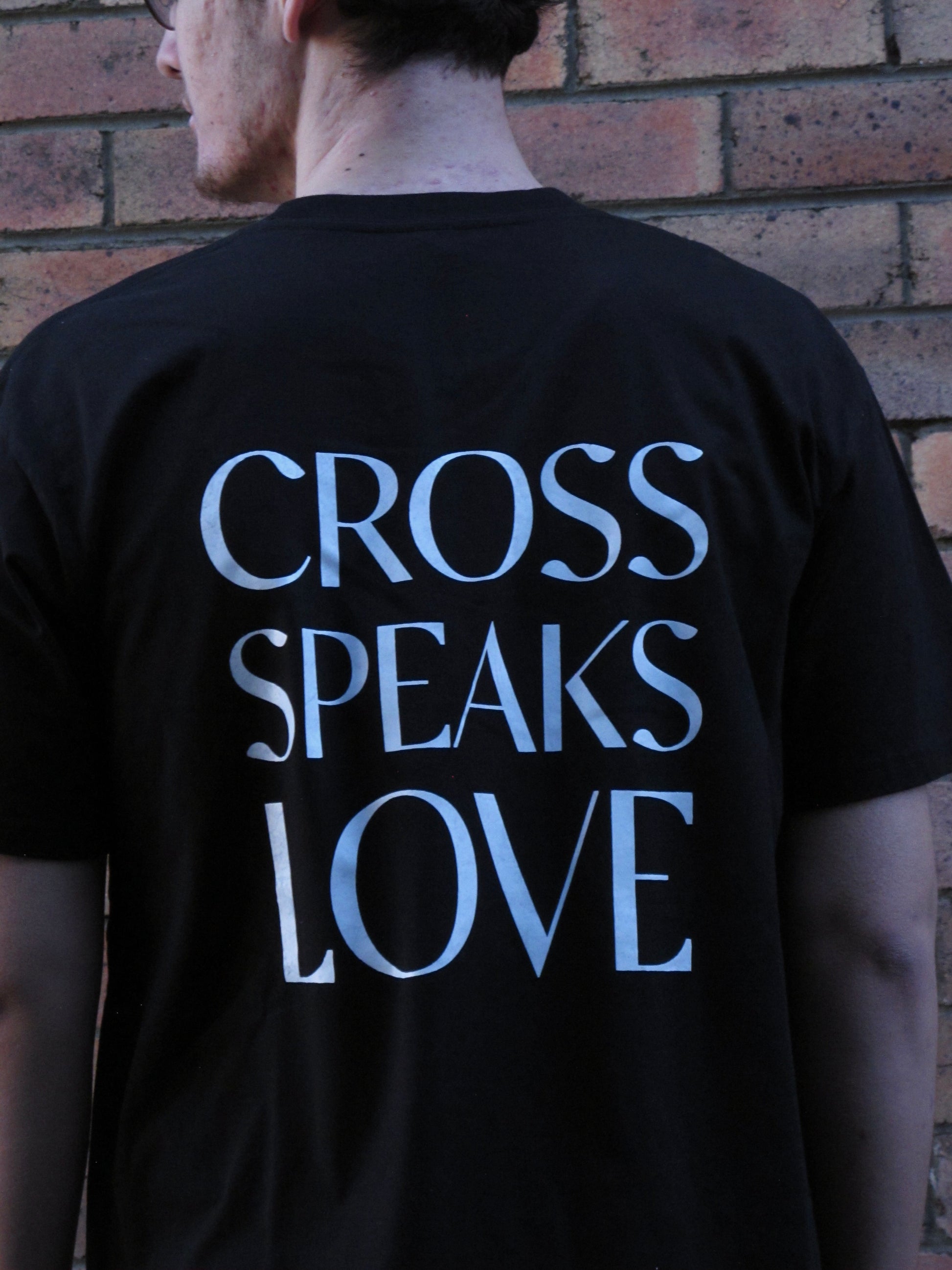 Male model wearing black tshirt with white ink design of words CROSS SPEAKS LOVE in capitals across his mid bacl. His head is turned to the left and a brick wall behind. 