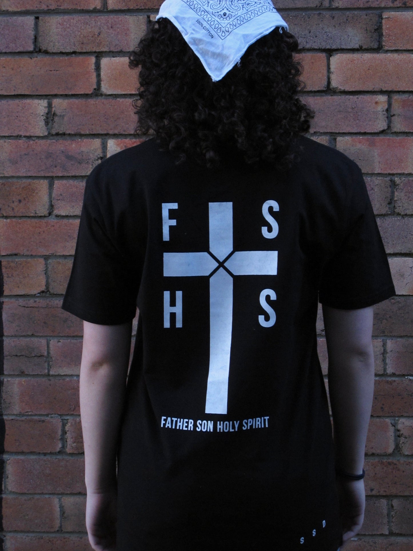 NEW DESIGN - Female model facing brick wall. FSHS in the four squares of the cross stating the first letters of the words Father Son Holy Spirit. Design aslo has large cross with rope like design in centre and the words FATHER SON HOLY SPIRIT written in capitals along the base. Right bottom side SSD acronym. All prints in white ink. on black shirt