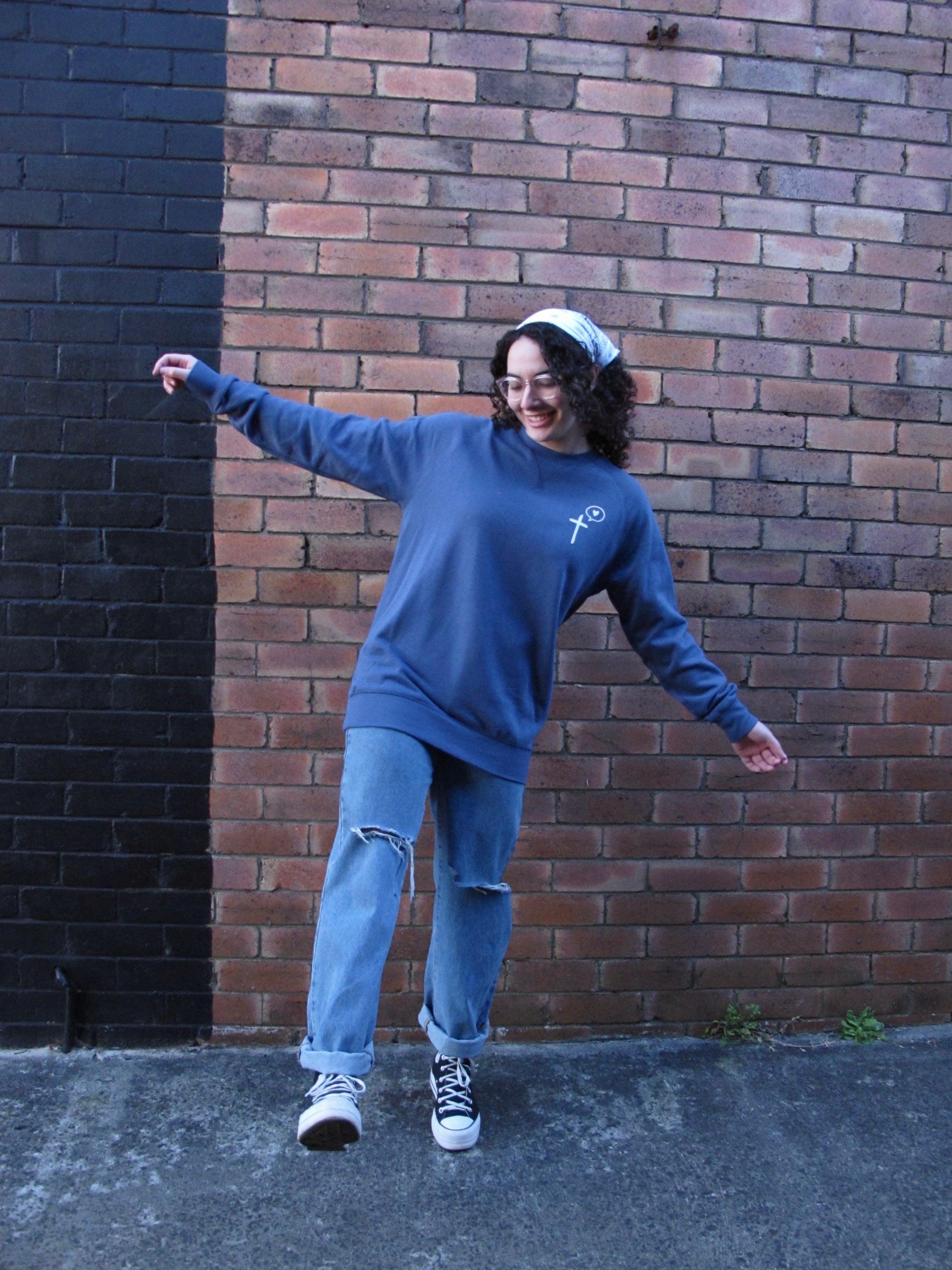 A fun pic of female model with white scarf in her hair, wearing glasses and arms outwide as she lifts one leg up as though dancing. She wears a diesel blue sweatshirt with left side pocket print. Model stand in front of a half black painted brick wall. She is smiling.