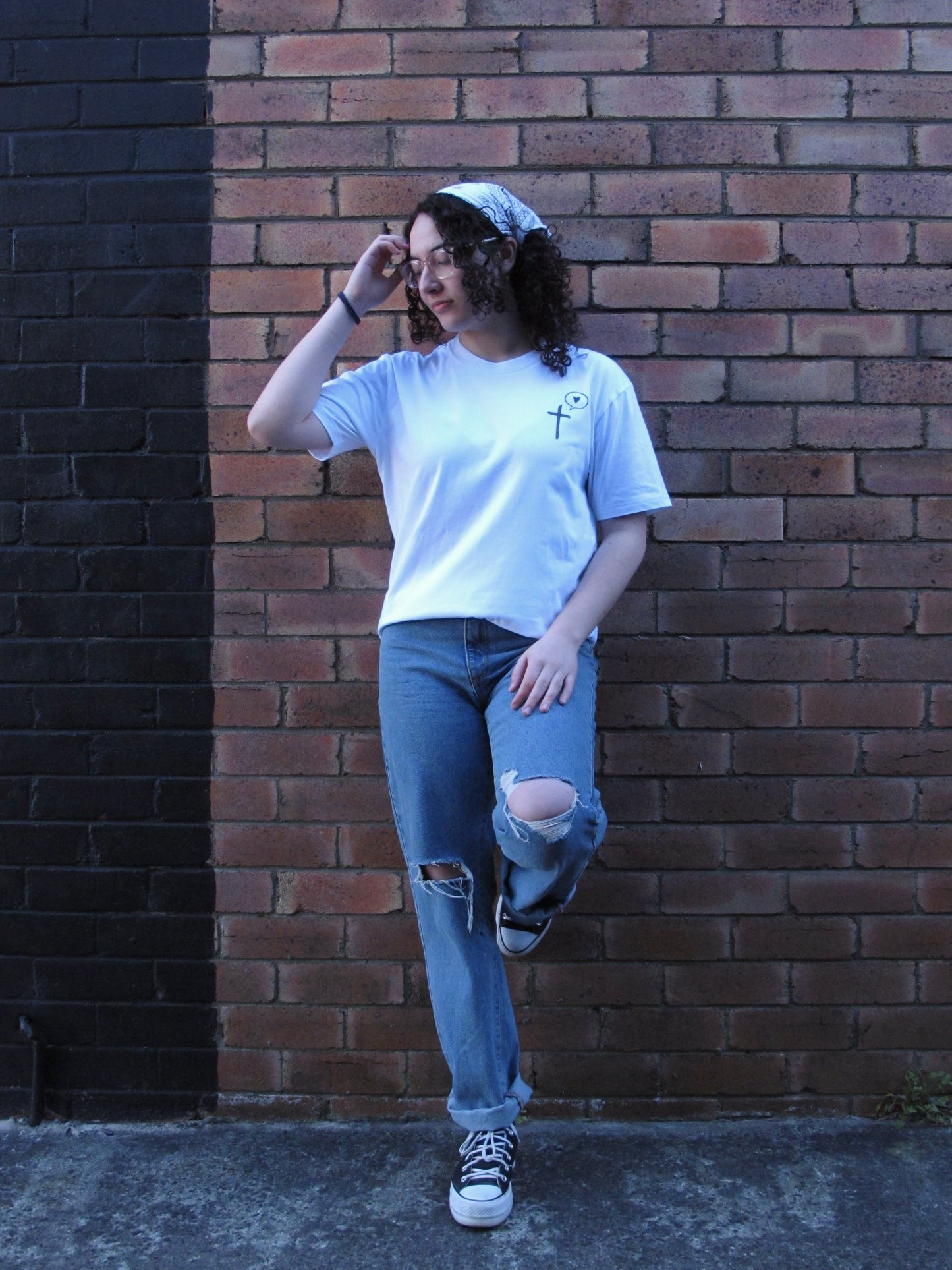 Young lady model - curly brown hair and white scarf in hair for style.  She is wearing a white CROSS SPEAKS LOVE tee and blue ripped jeans and converse shoes. She is leaning against a brick wall and looking down.