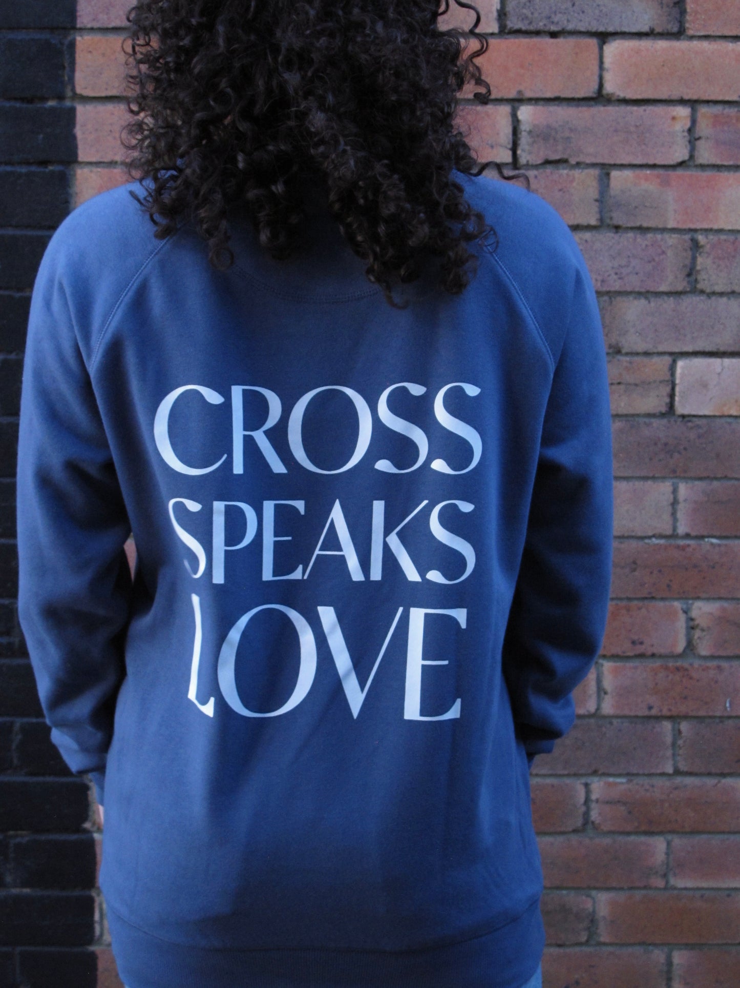 Female curly hair brunette standing facing a brick wall. She is wearing diesel blue sweatshirt with CROO SPEAKS LOVE in capitals as single lines. 