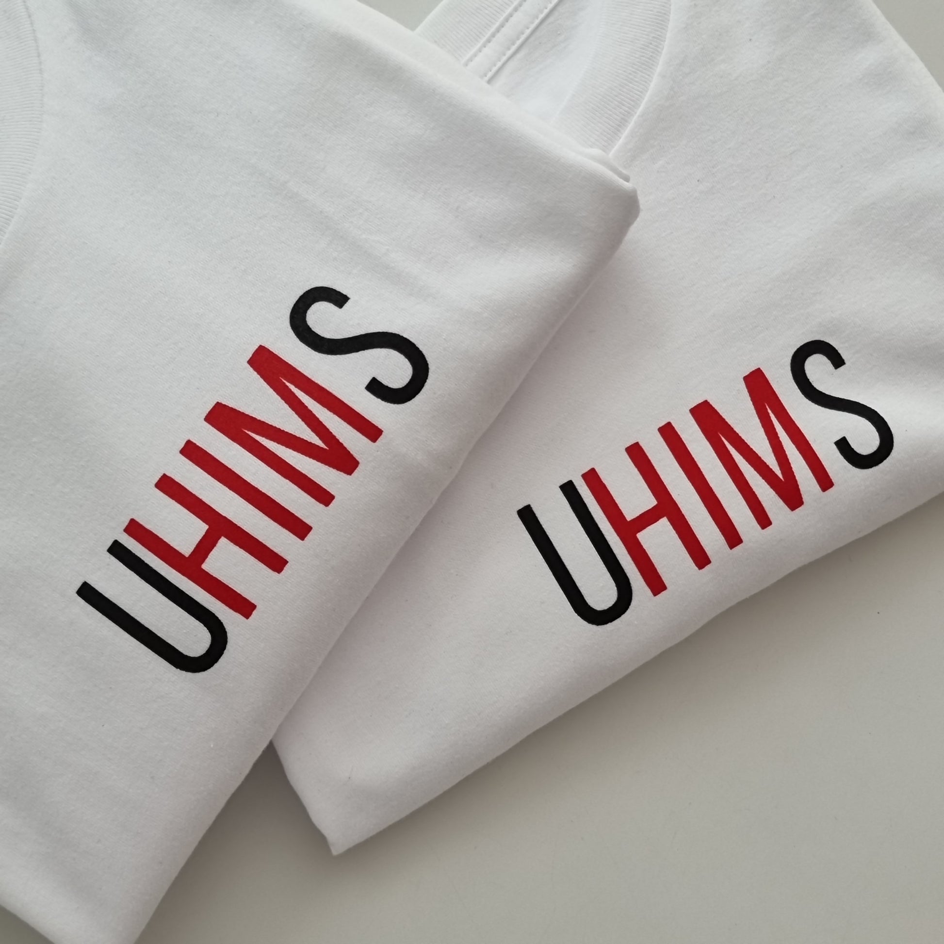 White tee with wordle like text. UHIMS written across front chest. Approximately 8 cms high and 20 cm wide across chest. U - Black ink, HIM - Red ink S - Black ink.