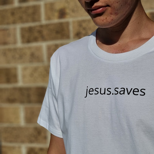 jesus.saves design written across chest in simple clear all lowercase font.  Model wears white tee with black ink text. Brick wall behind and he is looking down.  Print size is approx 2cm high and 20cm wide