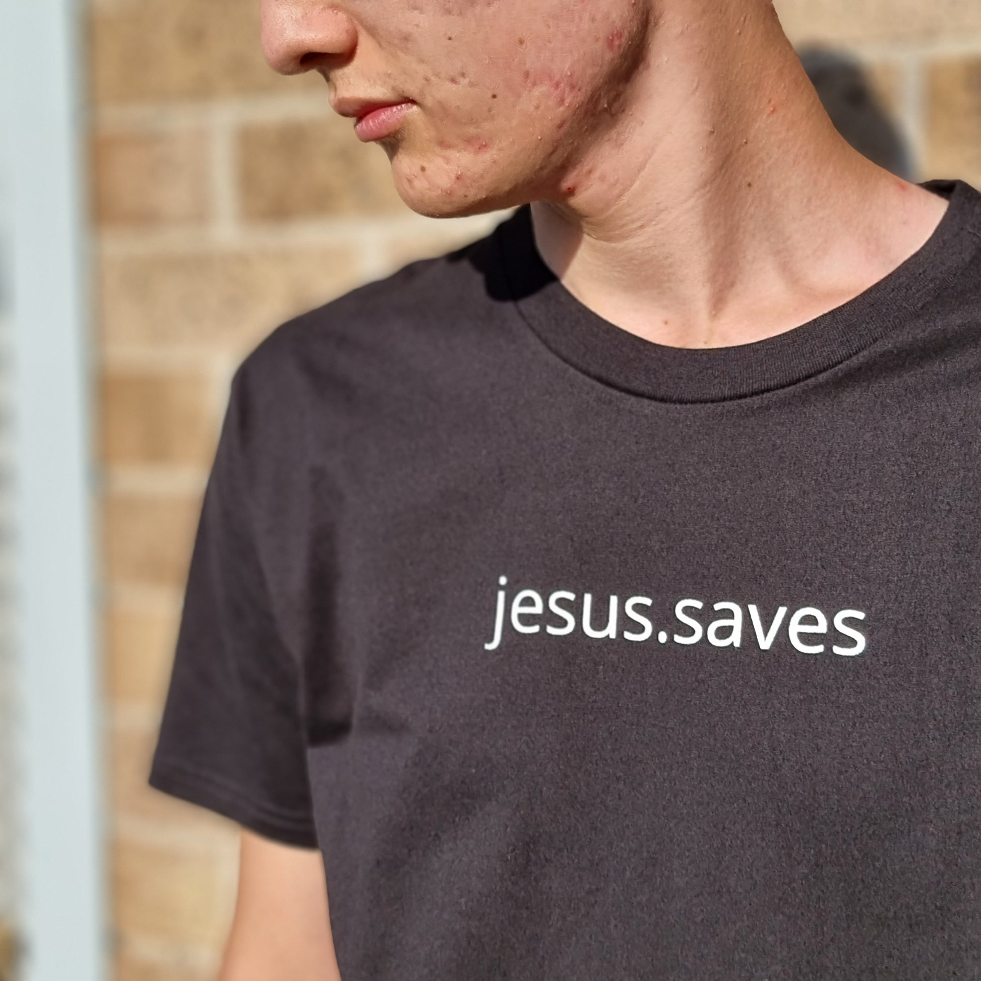 jesus.saves design written across chest in simple clear all lowercase font. Model wears black tee with white ink text. Brick wall behind and he is looking down. Print size is approx 2cm high and 20cm wide