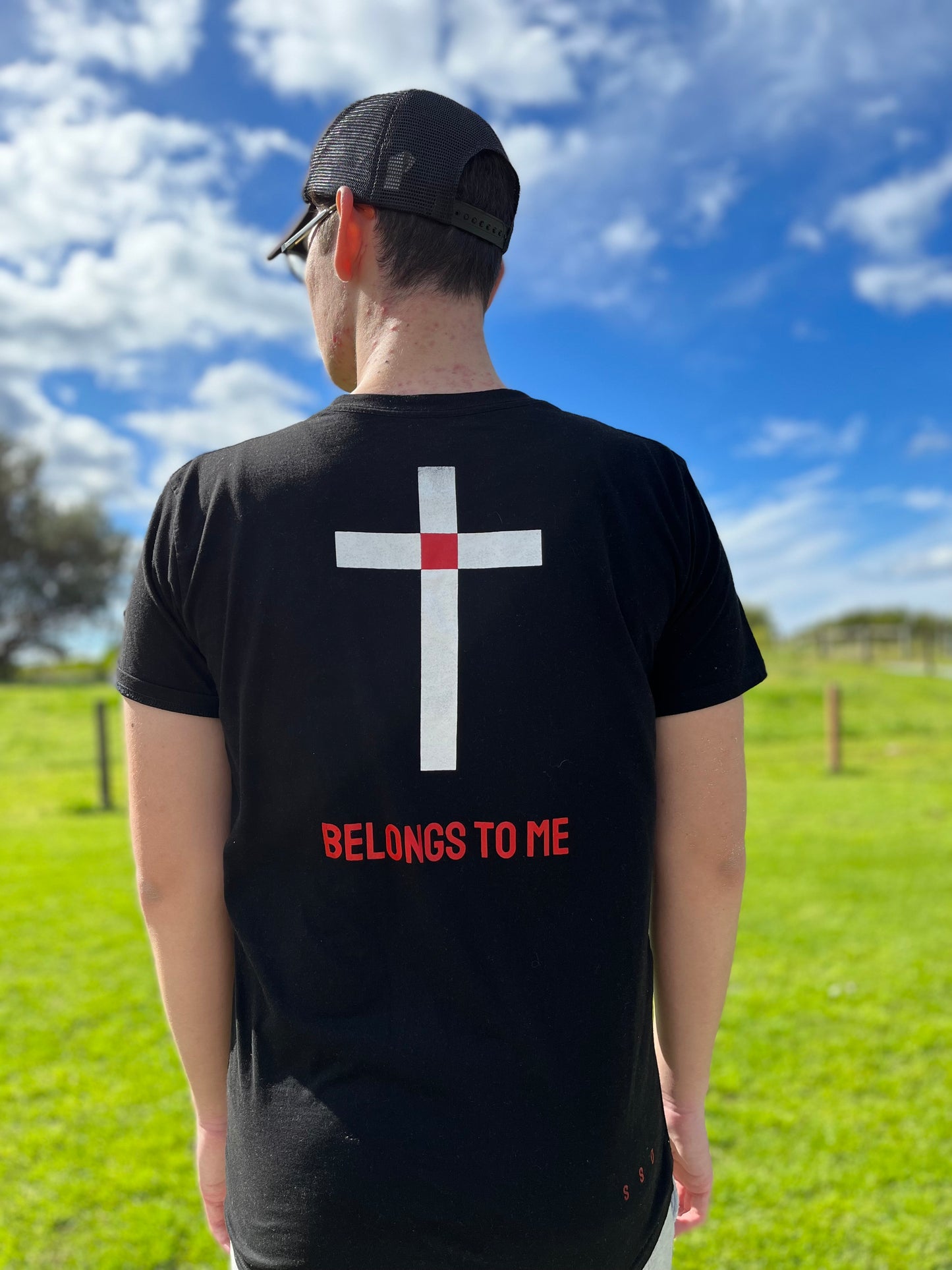 BELONGS TO ME tees. BLack. Blank front. Back design is white simple cross with red square at intersection point of cross that represents Christs blood shed on the cross. Red ink underneath in capitol letters states - BELONGS TO ME. Explaining that the cross is an identification symbol for Christians. Large back print. Long line shirt. Bottom right side SSD in red. Worn by model outside with blue sky and grass. 