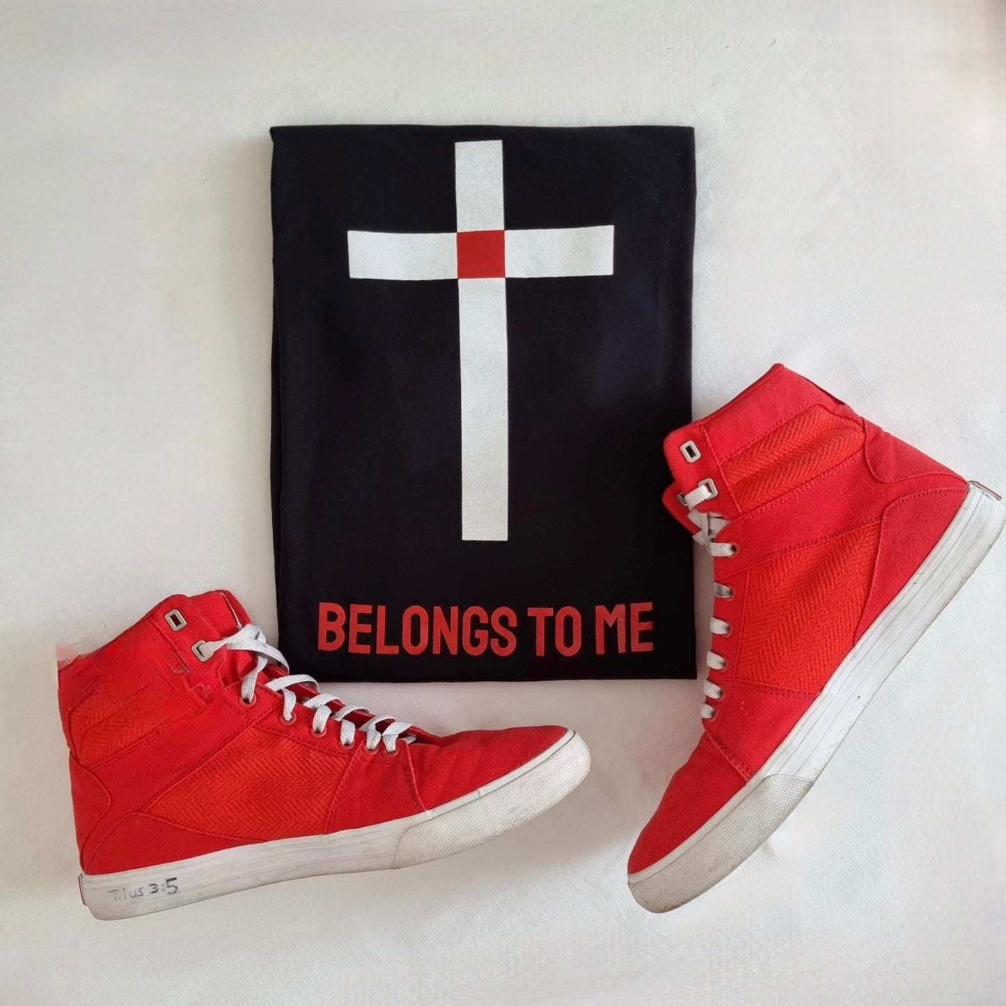 BELONGS TO ME tees. BLack. Blank front. Back design is white simple cross with red square at intersection point of cross that represents Christs blood shed on the cross. Red ink underneath in capitol letters states - BELONGS TO ME. Explaining that the cross is an identification symbol for Christians. Large back print. Long line shirt. Shown here with cool red hi top sneakers 